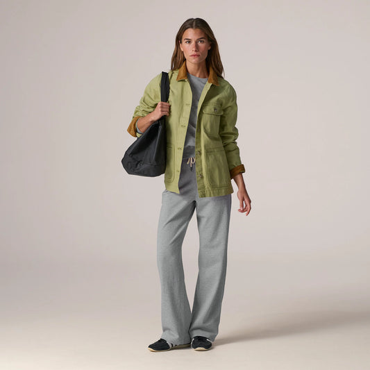 A woman stands against a plain light beige studio background wearing an olive green utility jacket with brown trim over a gray top and light gray wide-leg pants, while carrying a black shoulder bag.