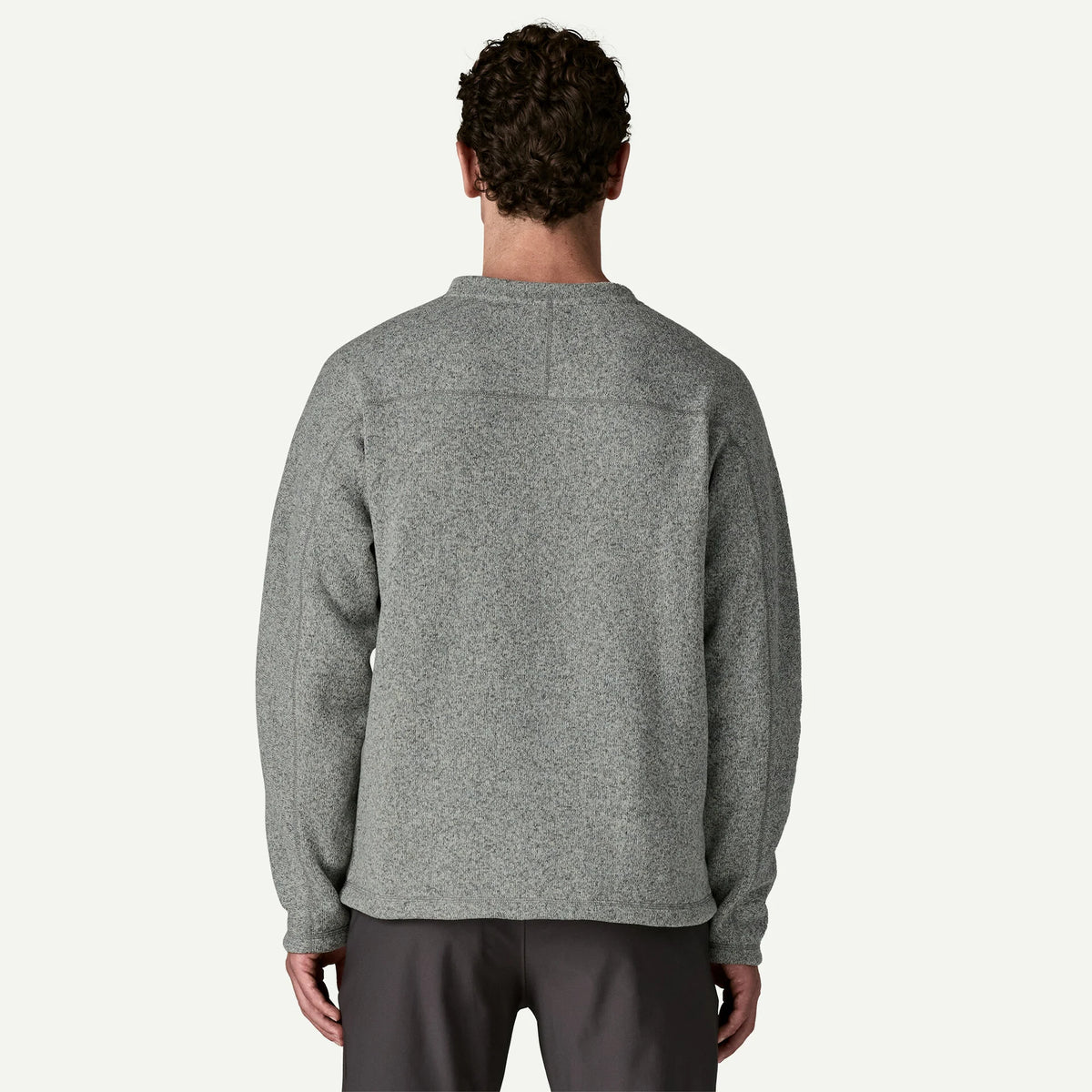 The back view of a man standing in a white studio wearing a grey sweater and dark pants