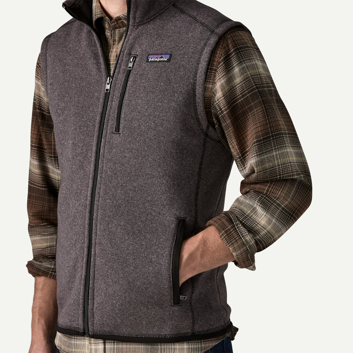 MAN STANDS WIHT HIS HADN I THE POCKET OF HIS grey BETTER SWEATER VEST.