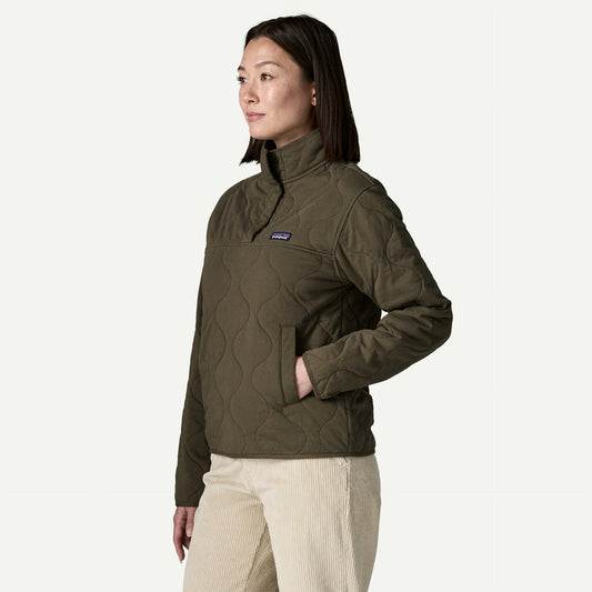 A woman stands in a white studio space wearing a quilted green sweater and cream coloured cords