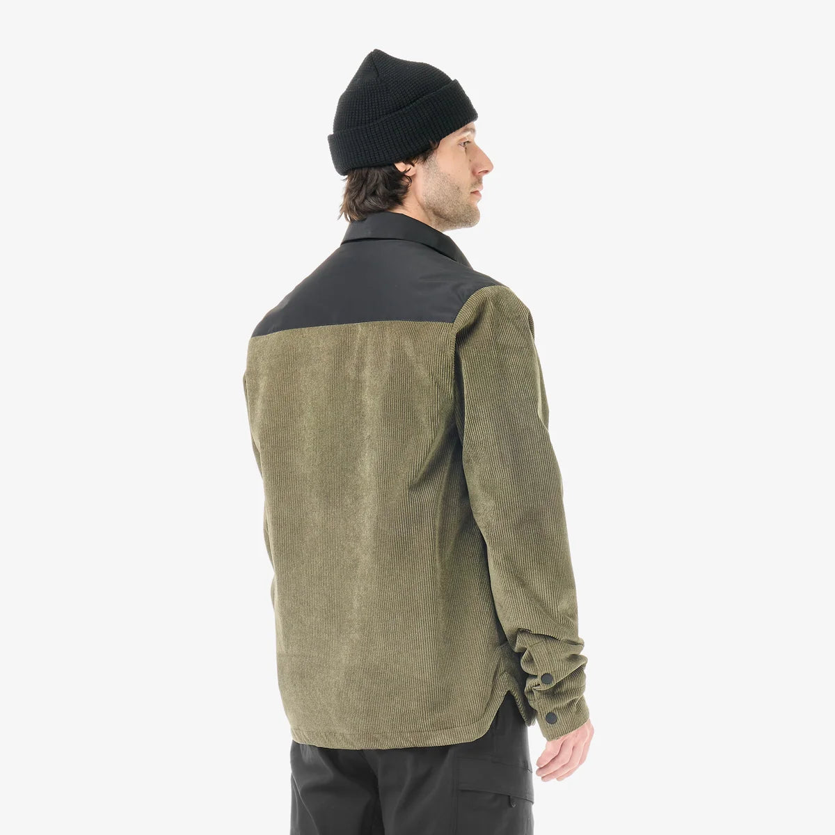 The back view of A man stands in a white studio wearing a green cord jacket with a black beanie and black pants