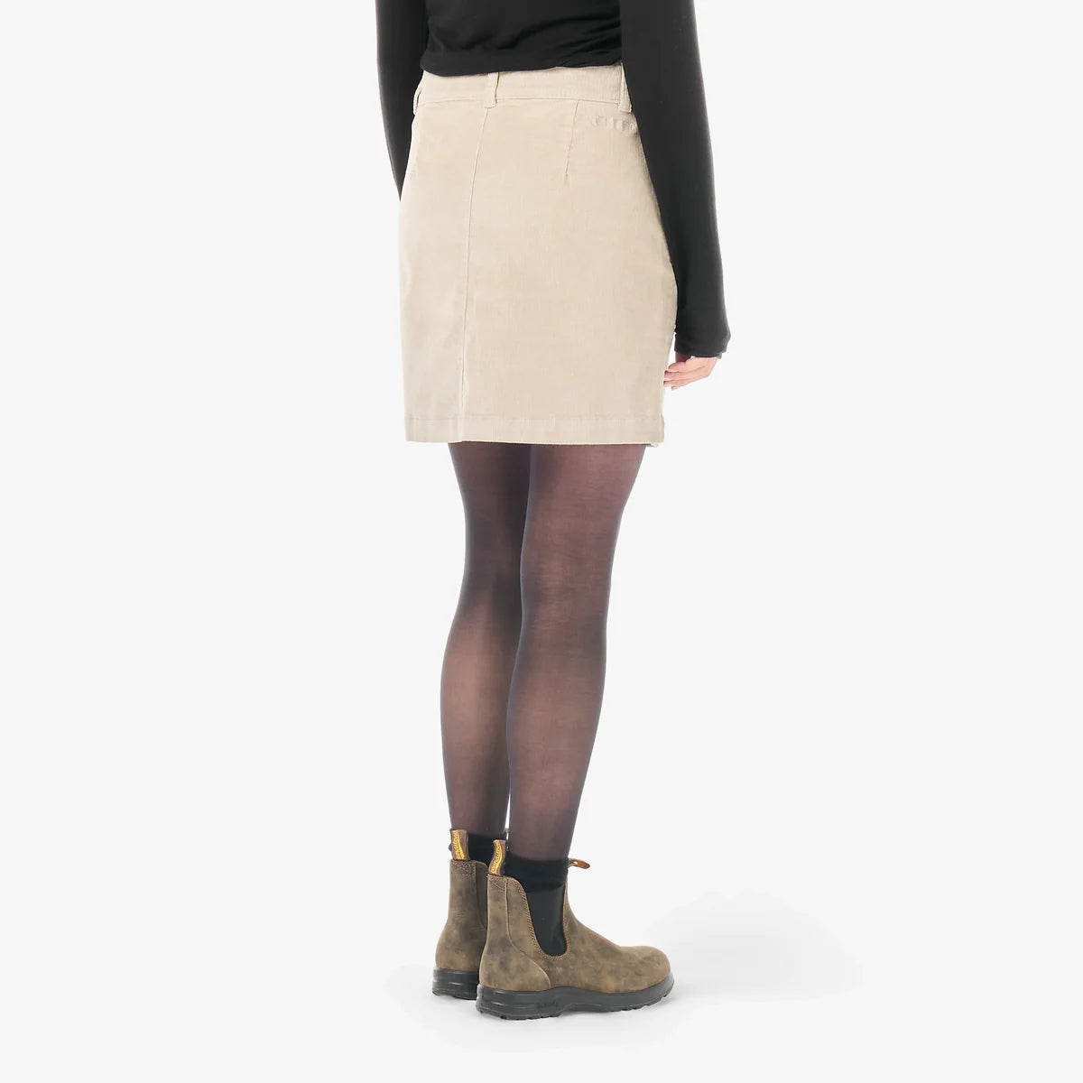 A woman stands in a white studio space wearing a cord mini skirt and black top and boots