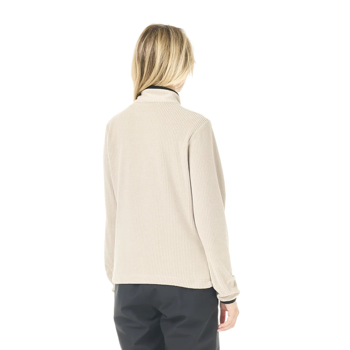 the back view of A woman stands in a white studio space wearing a cashmere coloured 1/4 zip top and black pants