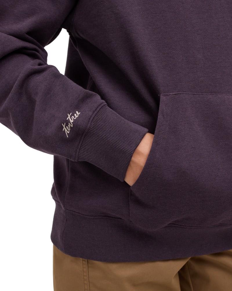 Tentree Field Foliage Hoodie Close-up of a person wearing a dark plum-purple pullover hoodie with a kangaroo pocket and a small white script logo on the left sleeve cuff, paired with tan pants against a white background.