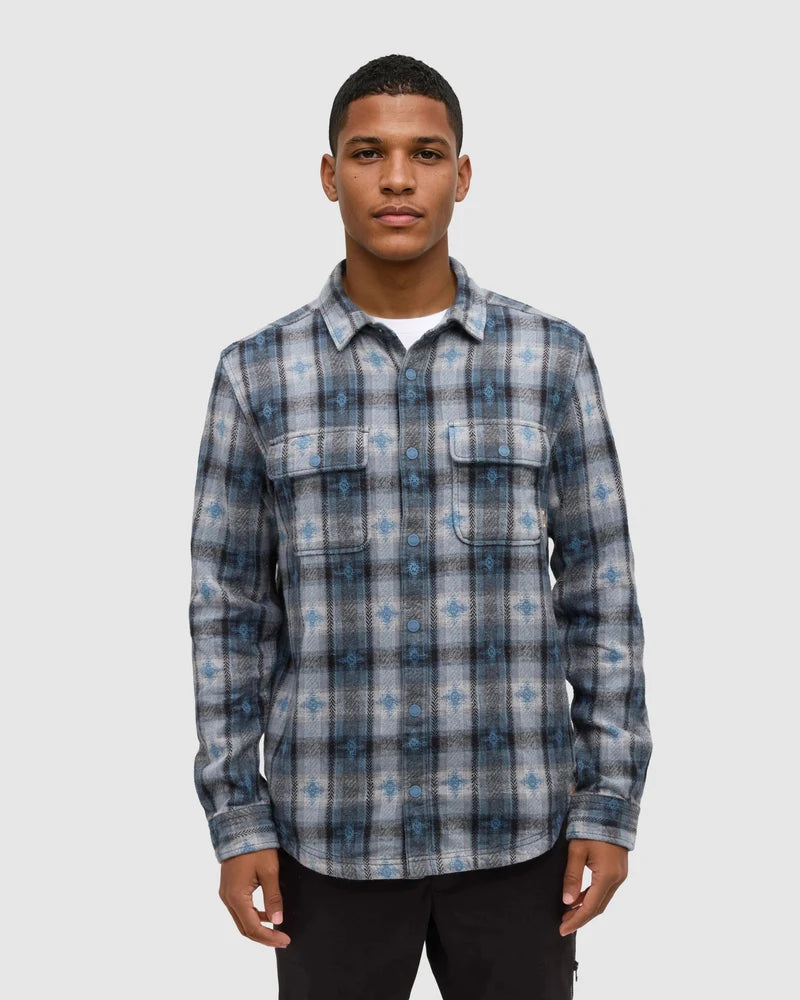 A man stands in a white studio space wearing a blue and grey button down shirt.