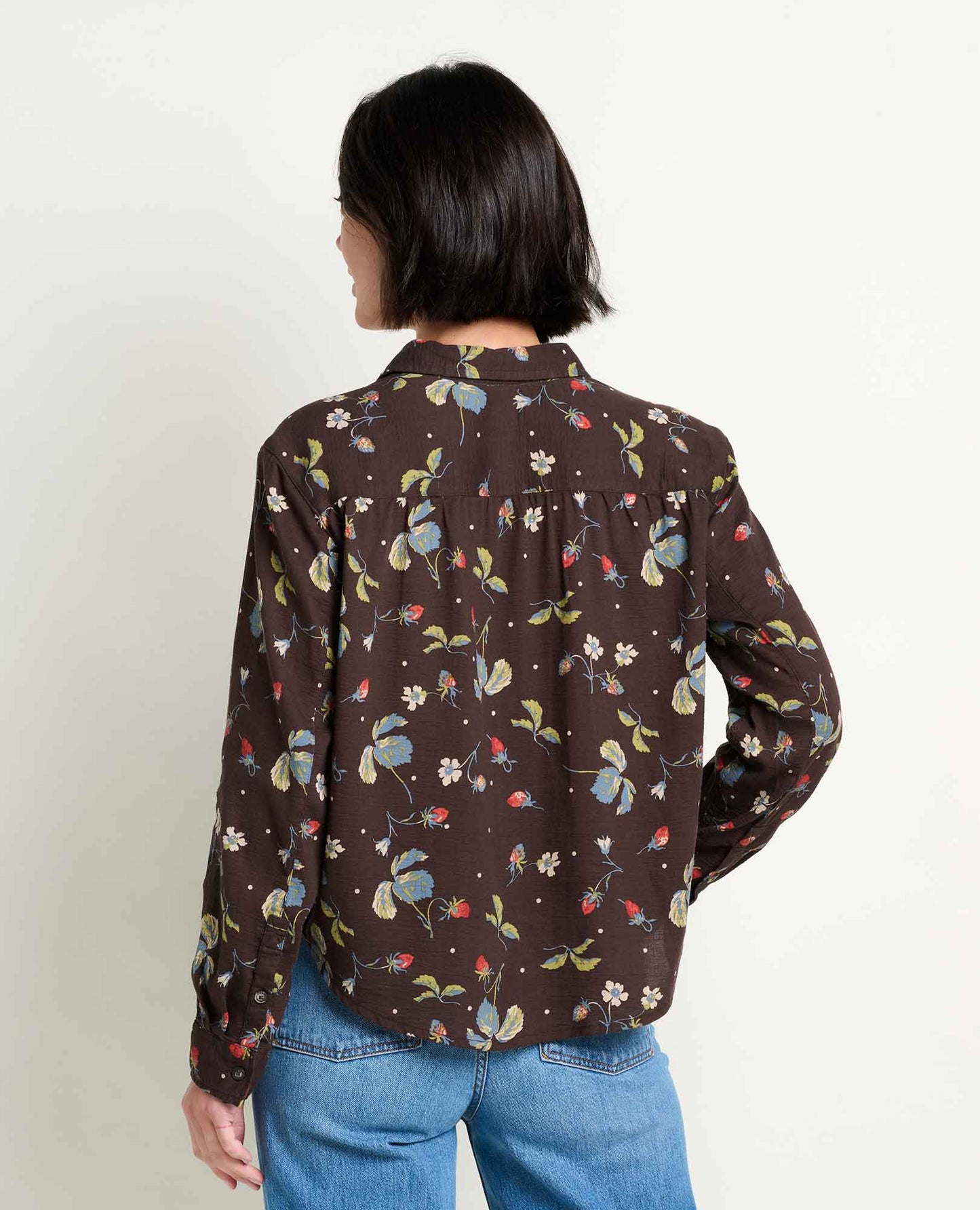 toad and CO manzana A woman viewed from behind wearing a dark brown long-sleeve button-up shirt with a colorful floral and butterfly print, paired with blue jeans, against a plain off-white background.