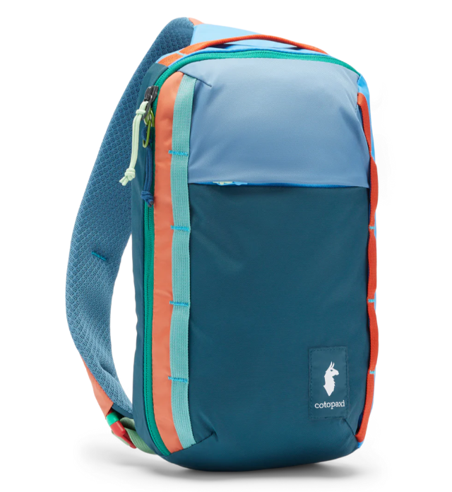 A blue and multicolored striped Cotopaxi Todo 8L Sling backpack with a zippered front pocket and a padded crossbody strap.