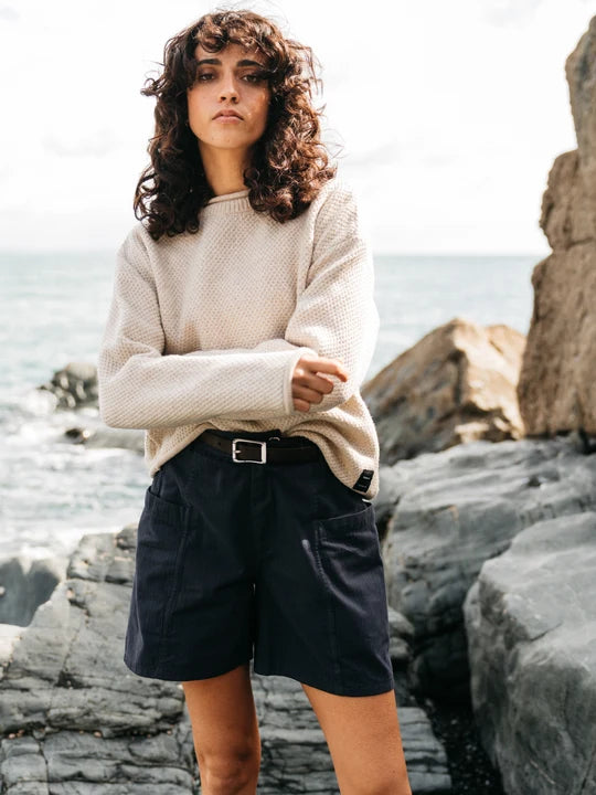 A woman standing beside the ocean wearing an oatmeal coloured wool sweater.