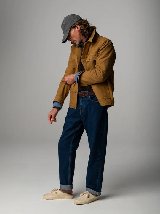 A make stands in a studio wearing a brown trucker jacket and blue jeans