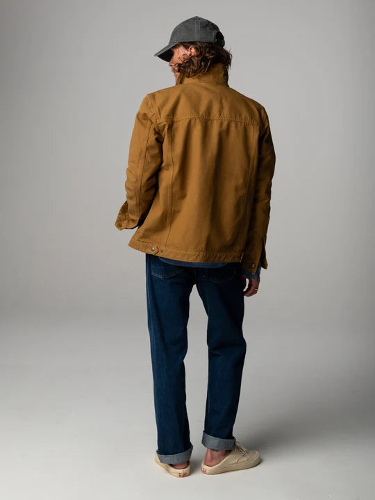 A rear view of a man standing in a studio space with grey background wearing a burnt ochre jacket and blue jeans