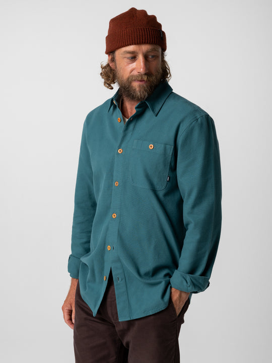 A man wearing a teal blue long-sleeve button-down shirt, a rust-brown beanie, and dark brown pants, standing against a light gray studio background.