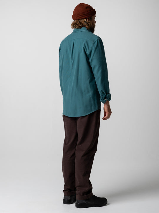 A man is shown from the back wearing a teal long-sleeve button-up shirt, dark brown pants, black shoes, and a rust-brown beanie against a light gray studio background.