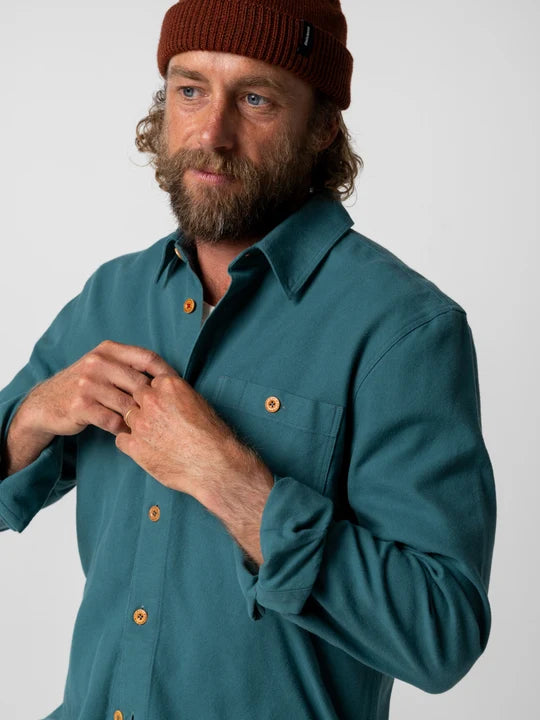 A man wearing a teal-green long-sleeve button-up shirt with orange buttons and a brown knit beanie, shown against a light gray studio background.