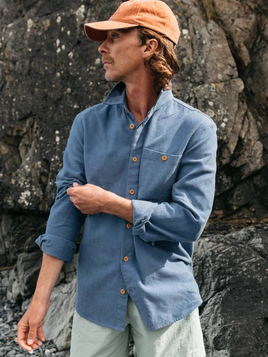 A man wearing a blue long-sleeve button-up shirt with brown buttons, an orange cap, and light green pants, standing against a dark rocky background.