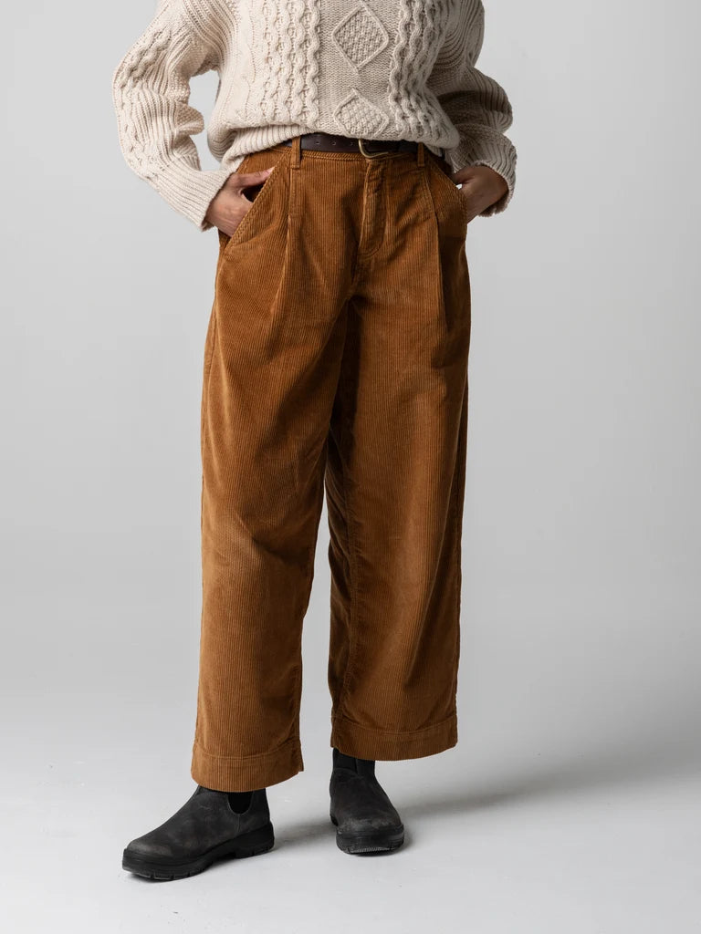 A person wearing brown corduroy wide-leg trousers, a cream cable-knit sweater, and black boots, standing against a light gray studio background.