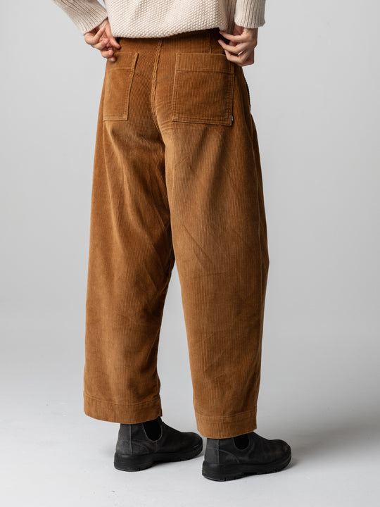A person shown from the back wearing loose brown corduroy pants, a cream knit sweater, and black slip-on shoes against a light gray studio background.