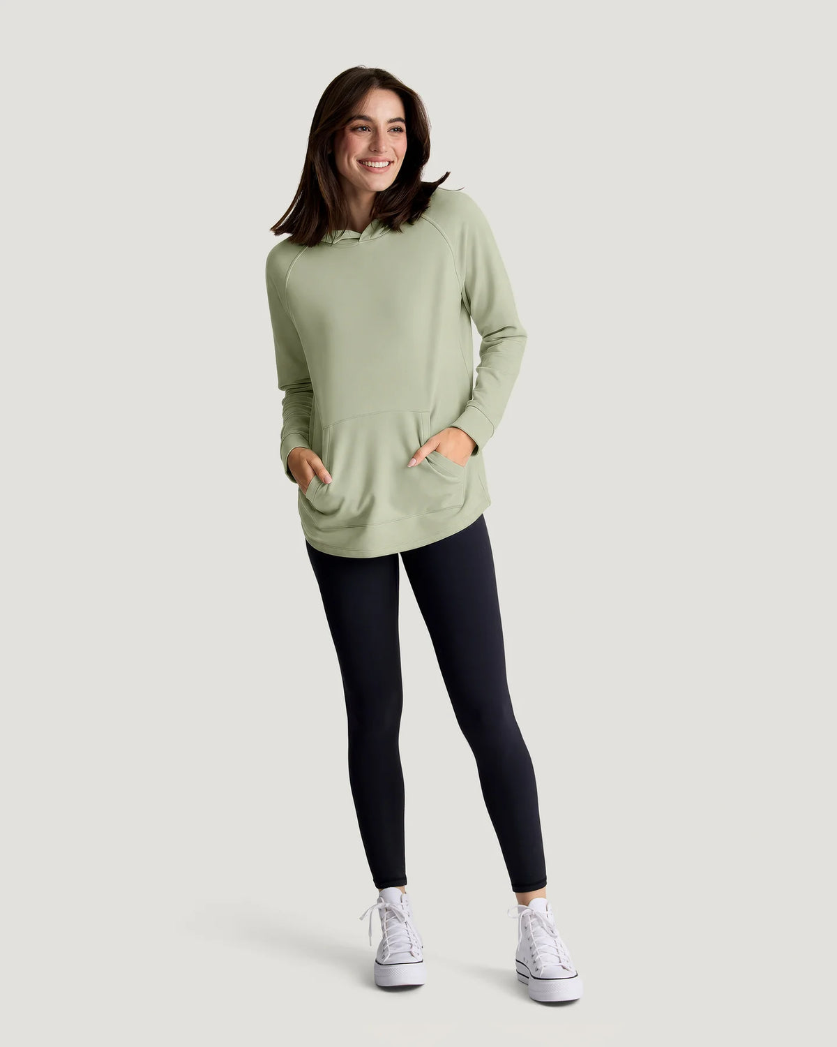 A woman standing in a studio with white walls wearing a green bamboo hoodie and tights