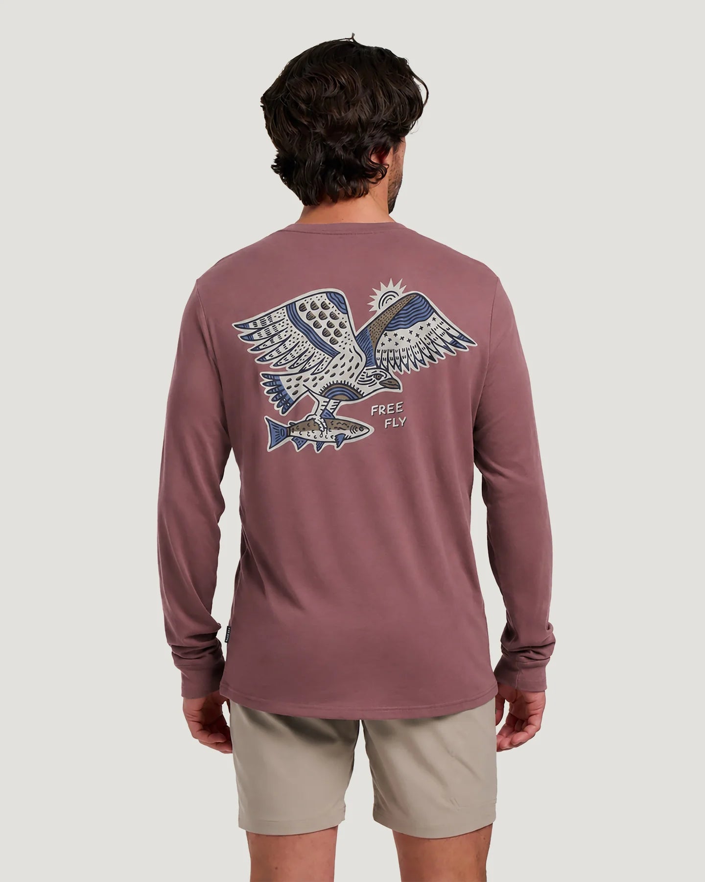 Free Fly Osprey Catch Long Sleeve A man viewed from behind wearing a dusty rose long-sleeve t-shirt with a large graphic of an eagle holding a fish on the back, paired with beige shorts, against a light gray background.