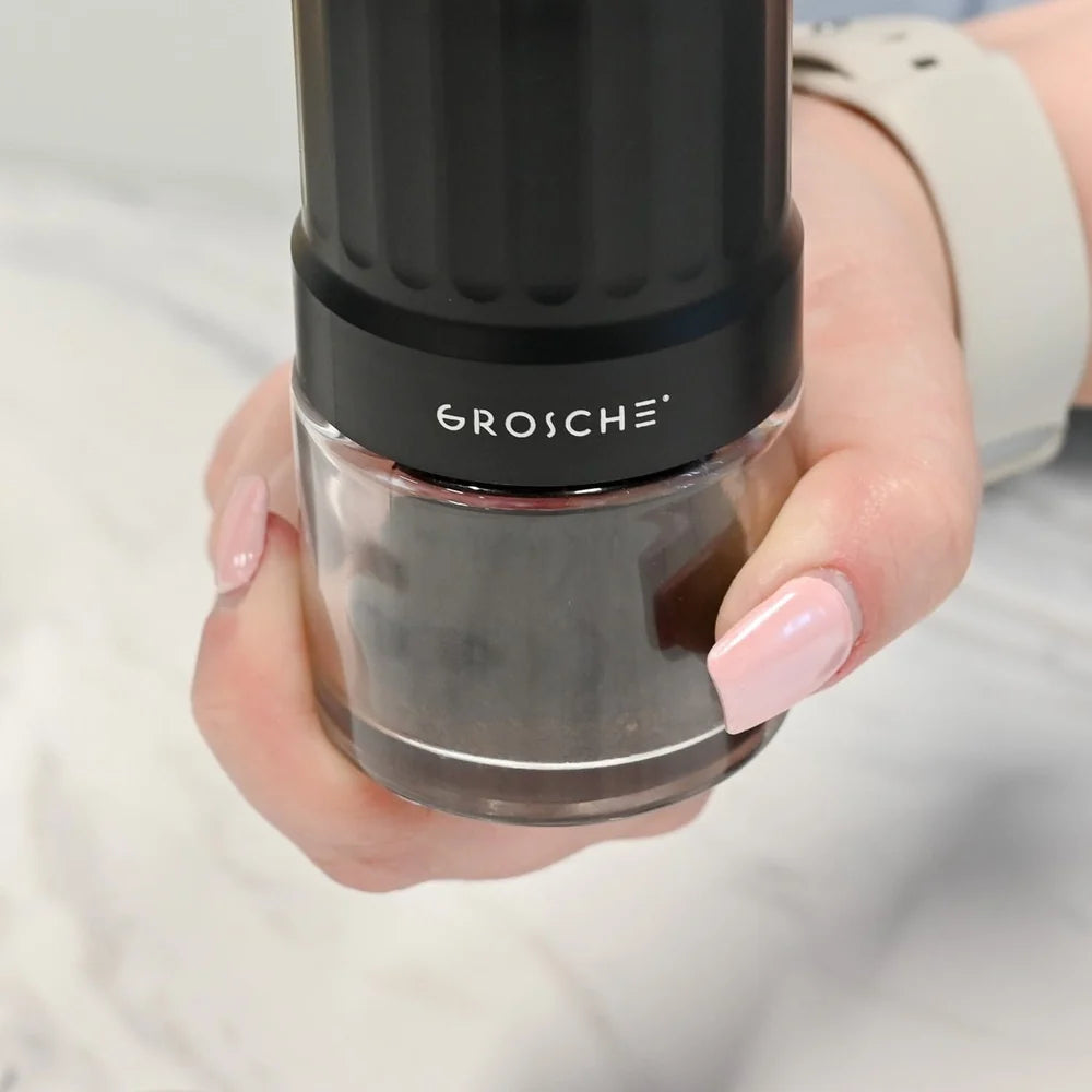 Grosche Tokio Manual Coffee Grinder A close-up of a hand holding a black and clear GROSCHE pepper mill grinder against a light marble background.