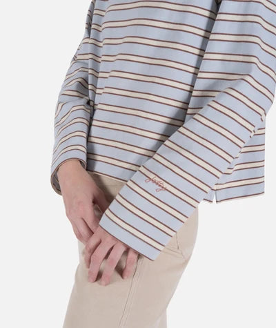 Jetty Hollis Striped Heavyweight Long Sleeve A close-up of a person wearing a light blue long-sleeve top with thin brown and beige horizontal stripes, paired with beige pants. The brand name 'jeffrey' is embroidered in brown on the cuff.