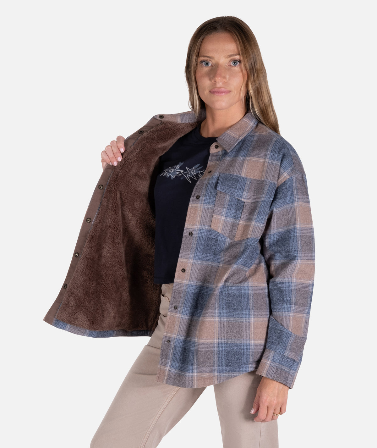 A woman stand in a studio with a blue and brown jacket open and cream pants
