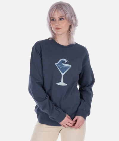A model with short light purple hair wearing a dark navy long-sleeve crewneck sweatshirt with a blue and white martini glass graphic on the chest, paired with beige pants, against a light gray background.