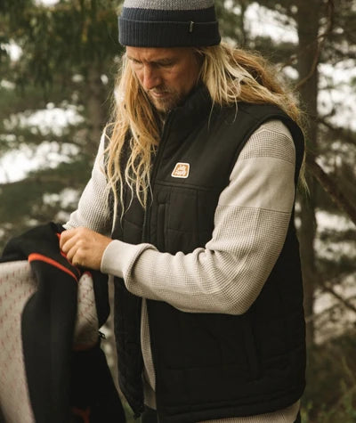 Jetty Terrace Vest A man with long blonde hair and a beard wearing a black quilted vest over a beige waffle-knit sweater and a dark beanie, standing outdoors in a snowy forest setting while holding a jacket.