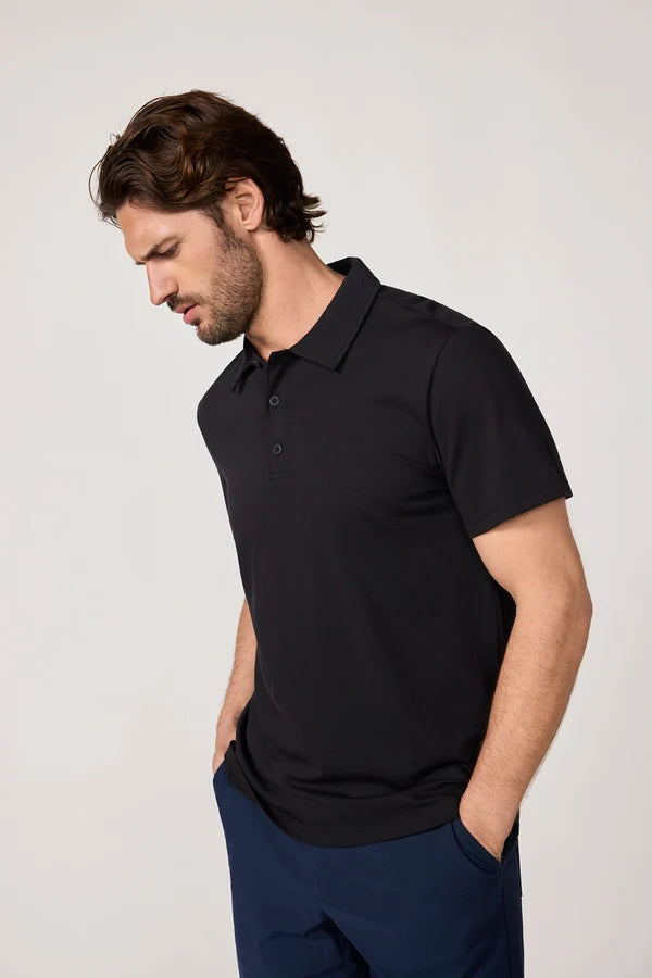 MPG Revive Short Sleeve Polo A man wearing a black short-sleeve polo shirt and navy blue trousers, standing against a light gray studio background.