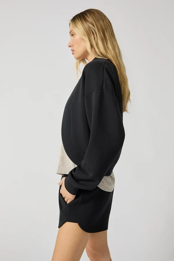 The side view of a woman that stands in a white studio space wearing a black cardigan shrug and black shorts