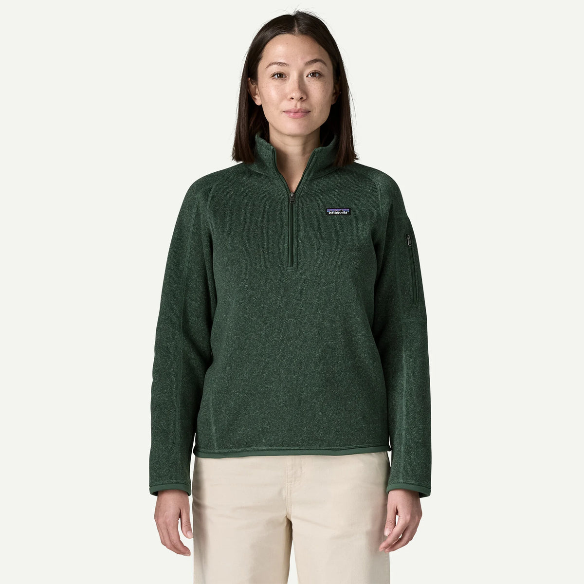 A woman stands in a whit studio wearing a 1/4 zip green better sweater and cream pants