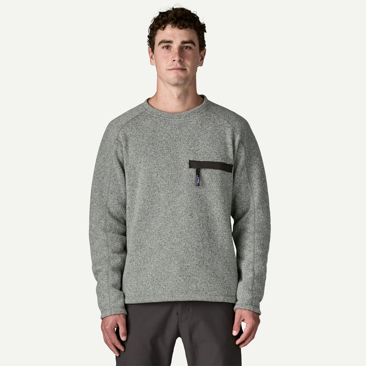 A man stands in room with white background wearing a grey sweater with front pocket with zipper.