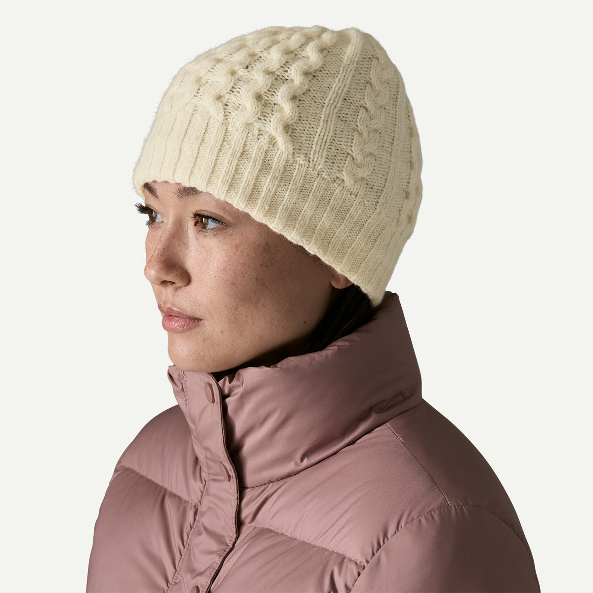 A woman wears a white cable knit beanie with a taupe winter jacket on a white background