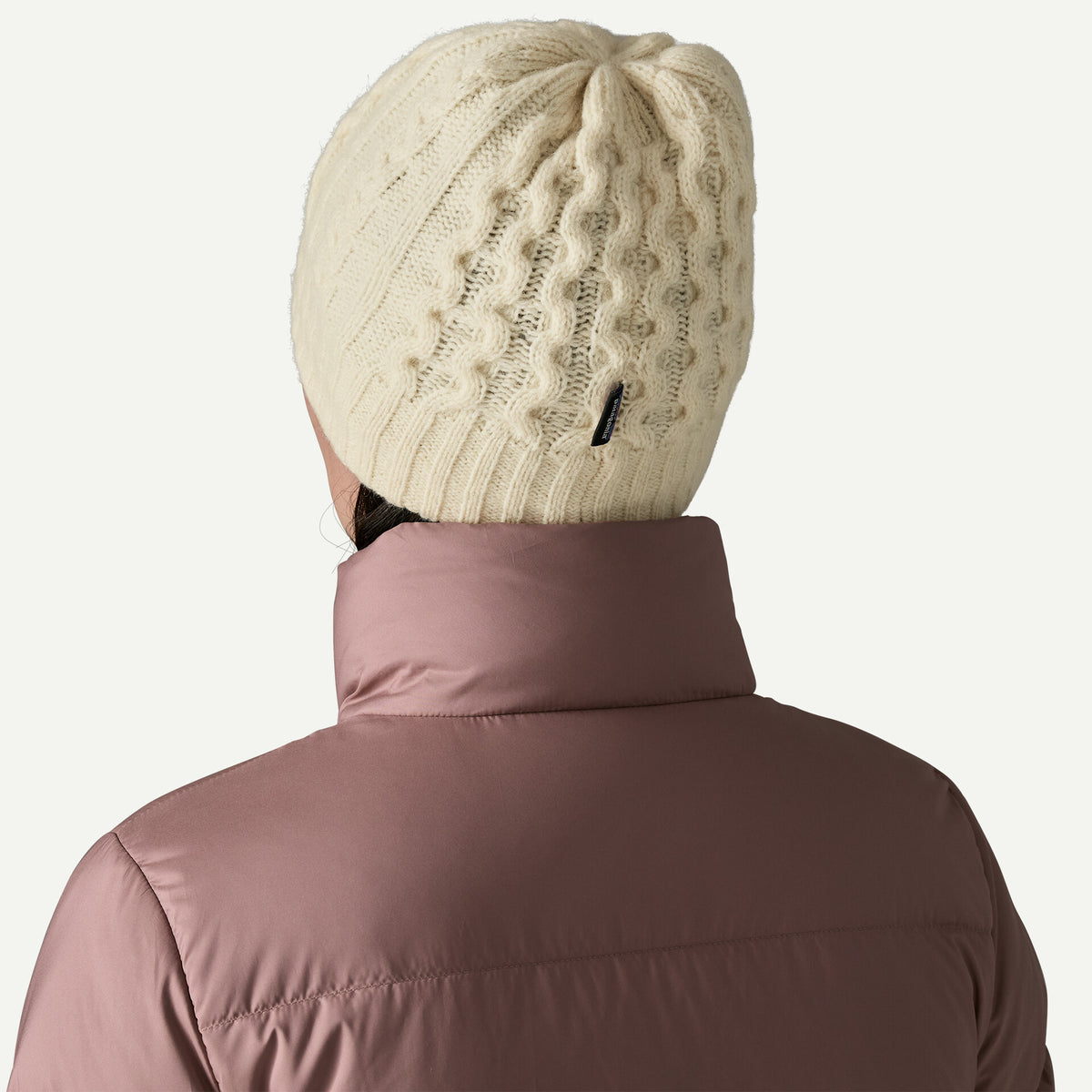 The back view of a woman wearing a white cable knit beanie and a taupe jacket in a white room