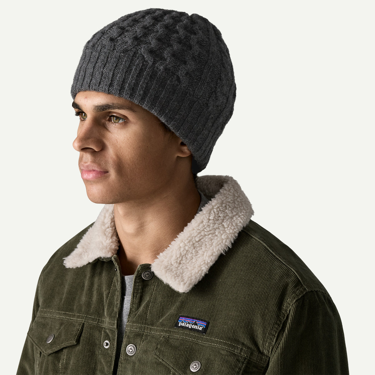 A man wears a grey cable knit beanie and a green cord jacket with a fleece collar