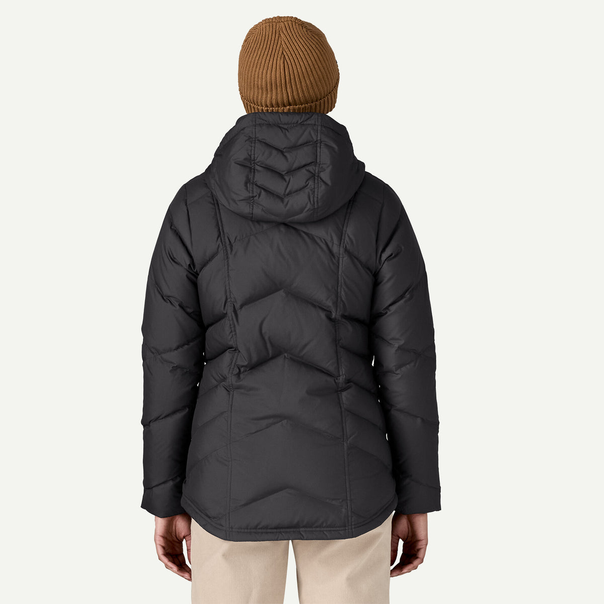 back view of a woman standing in white studio wearing black down jacket with a hood. She has crema pants on and a brown toque / beanie. 