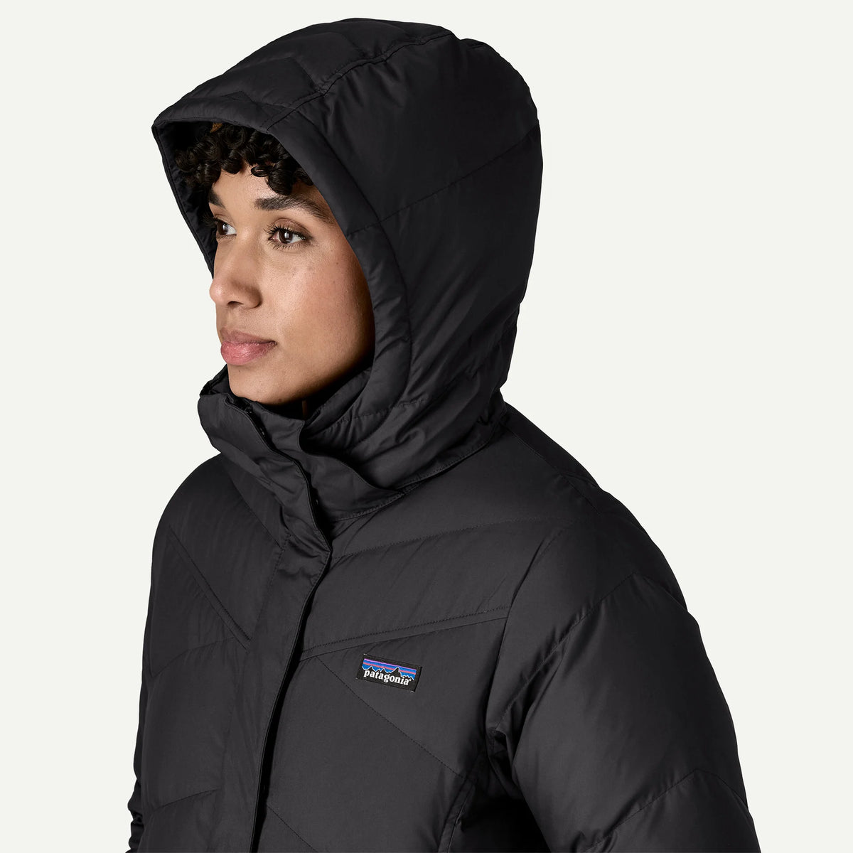 A woman stands with a white background wearing a black hooded down jacket