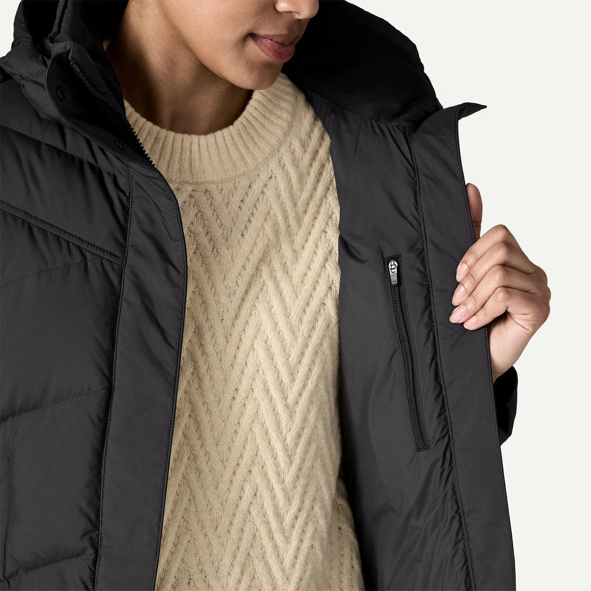 A woman shows the inside zip of her black down jacket with a cream sweater underneath.