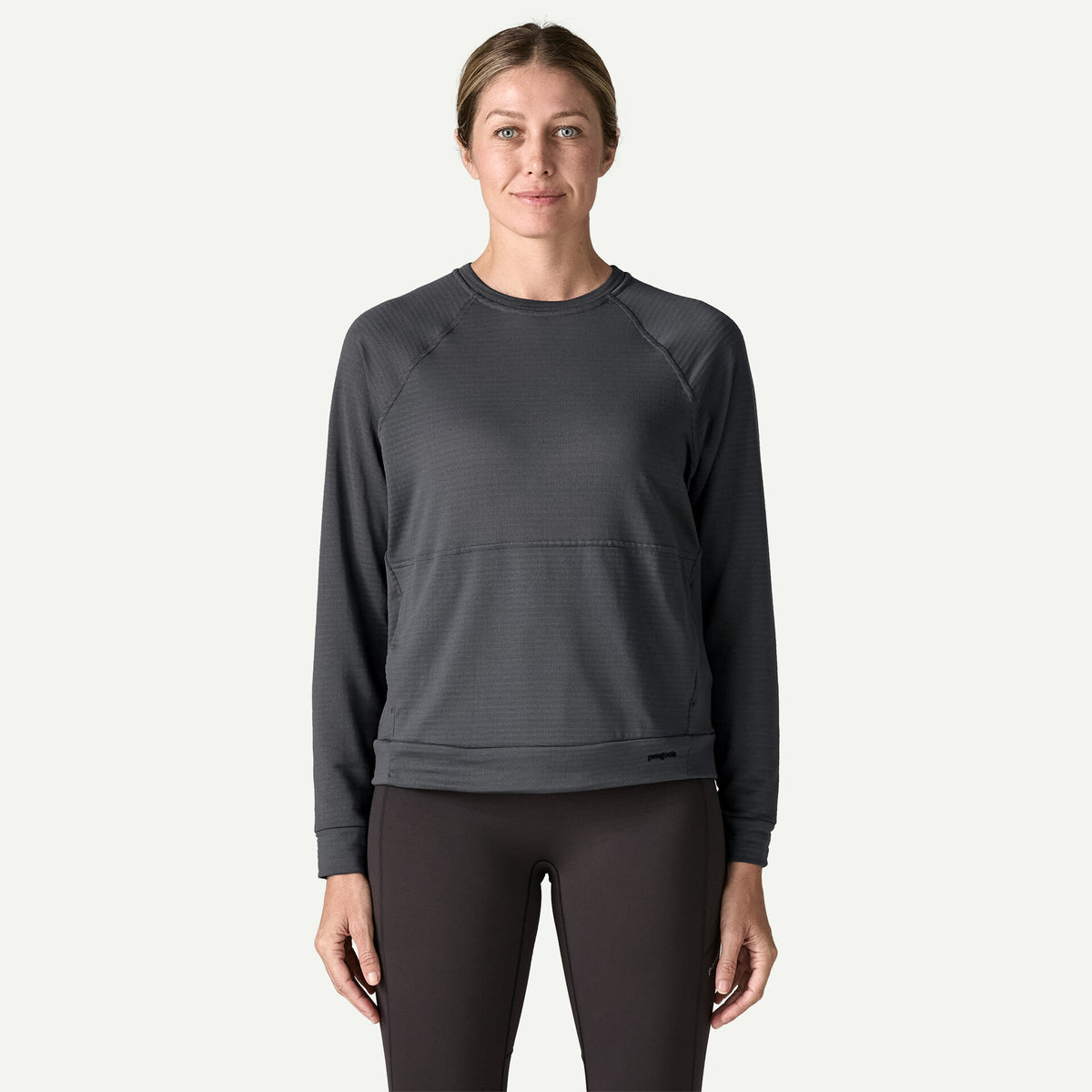 A woman stands in a white studio wearing a black crew with a front pouch pocket and black tights