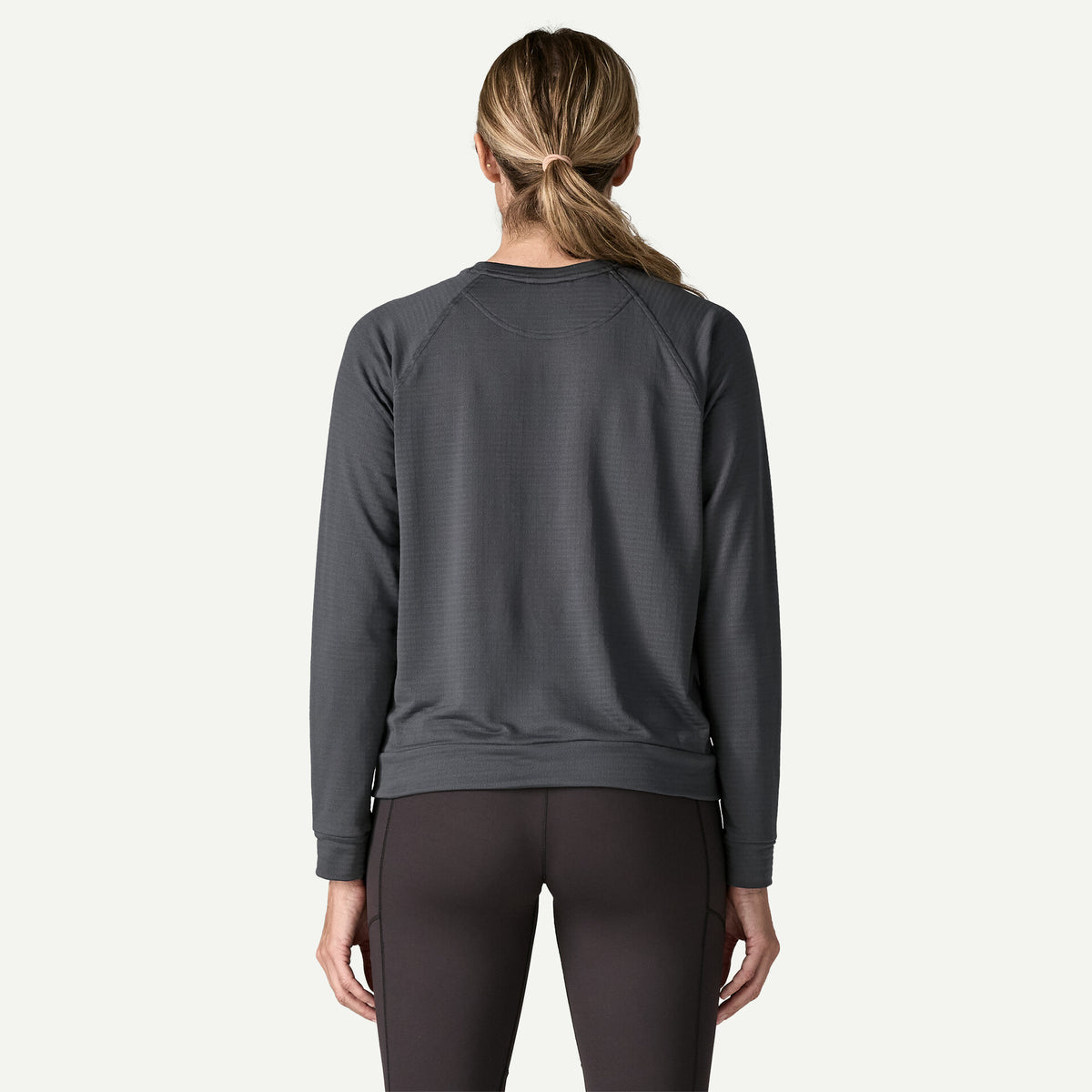 The back view of a woman standing in a white room wearing a black crew and black tights