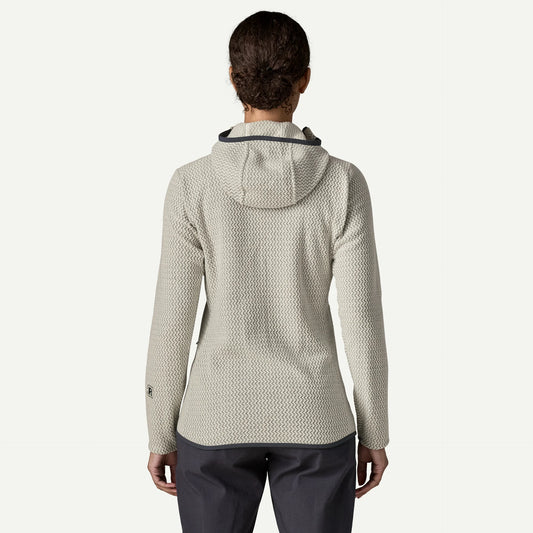The back view of a woman standing in a whit studio space wearing a hooded sweater and blue pants