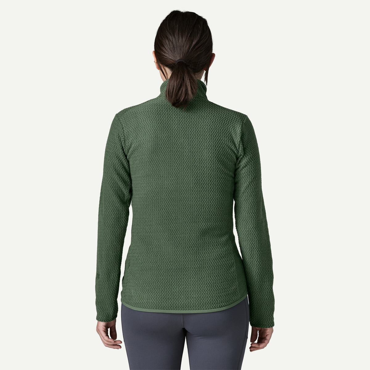 The back view of a woman standing in a white room wearing a green sweater and blue pants