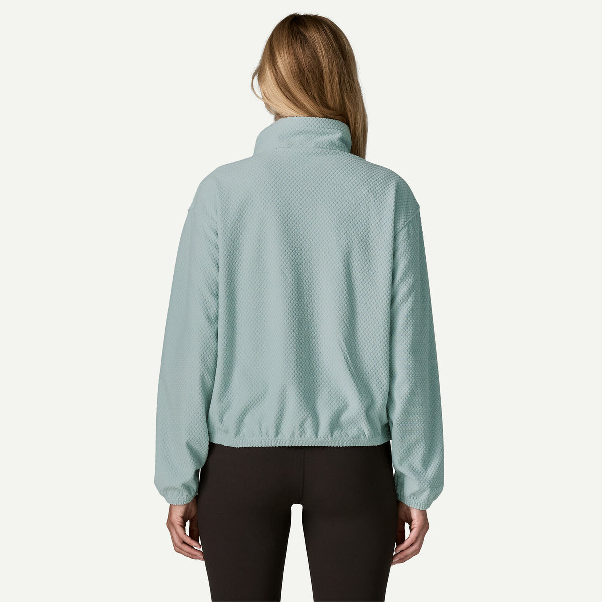 The back view of a woman standing in a studio space wearing a blue textured fleece and black pants