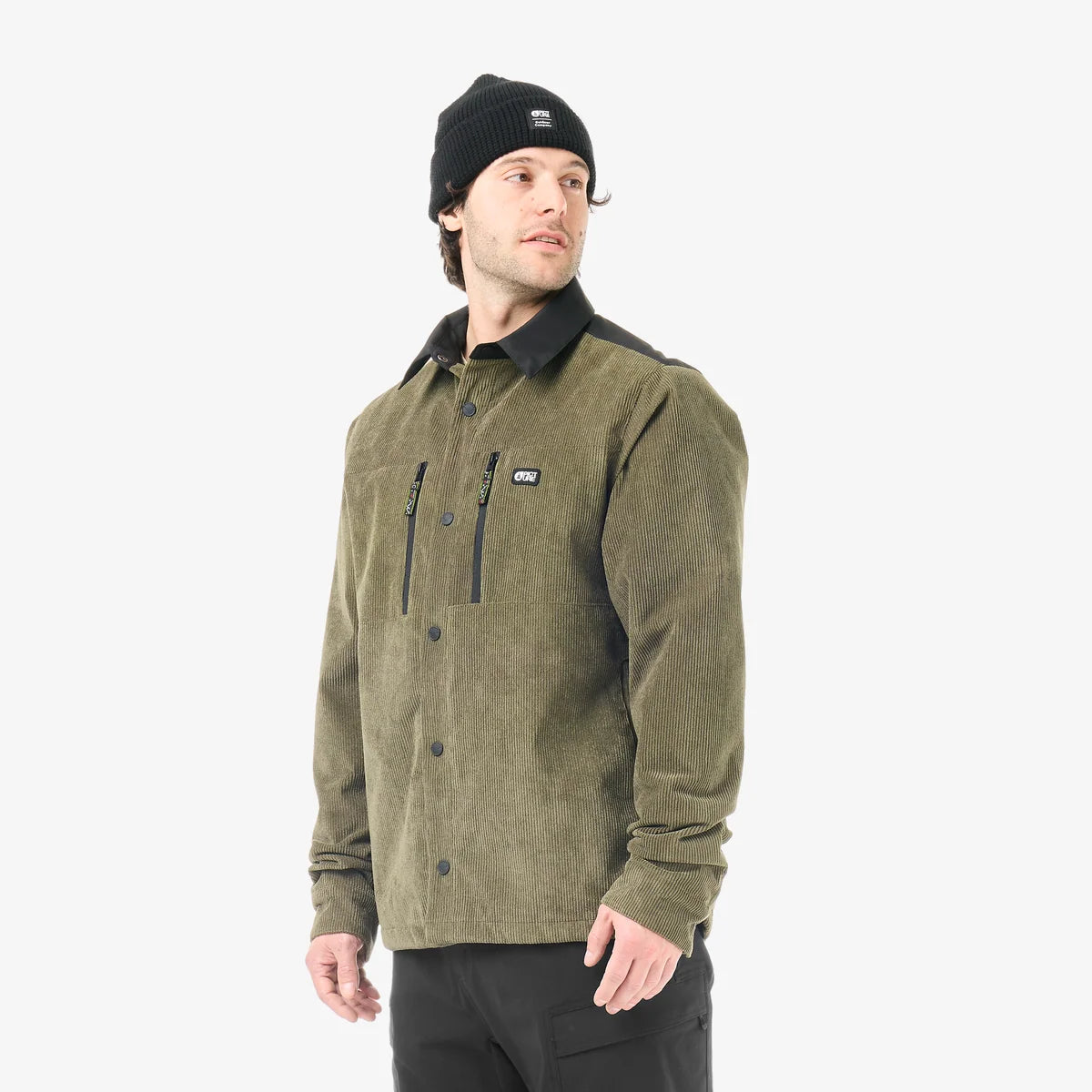 A man stands in a white studio wearing a green cord jacket with a black beanie and black pants