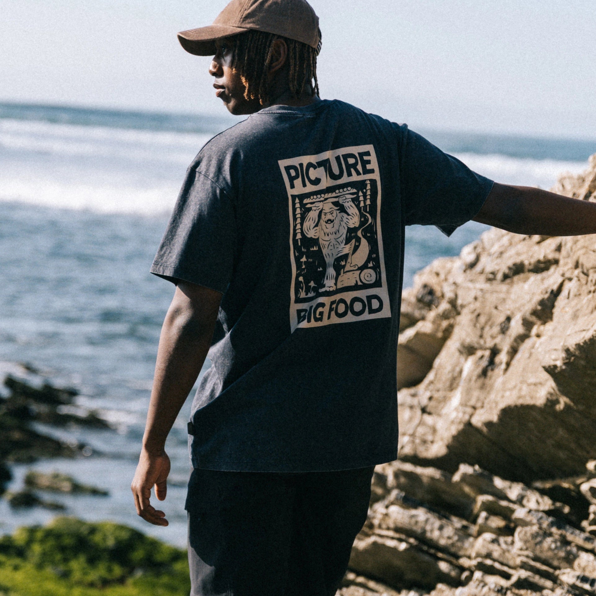 Picture Organic Big Foot Tee A man standing on a rocky beach wearing a dark charcoal gray t-shirt with a large white graphic print on the back that reads 'PICTURE BIG FOOD', paired with a brown baseball cap.