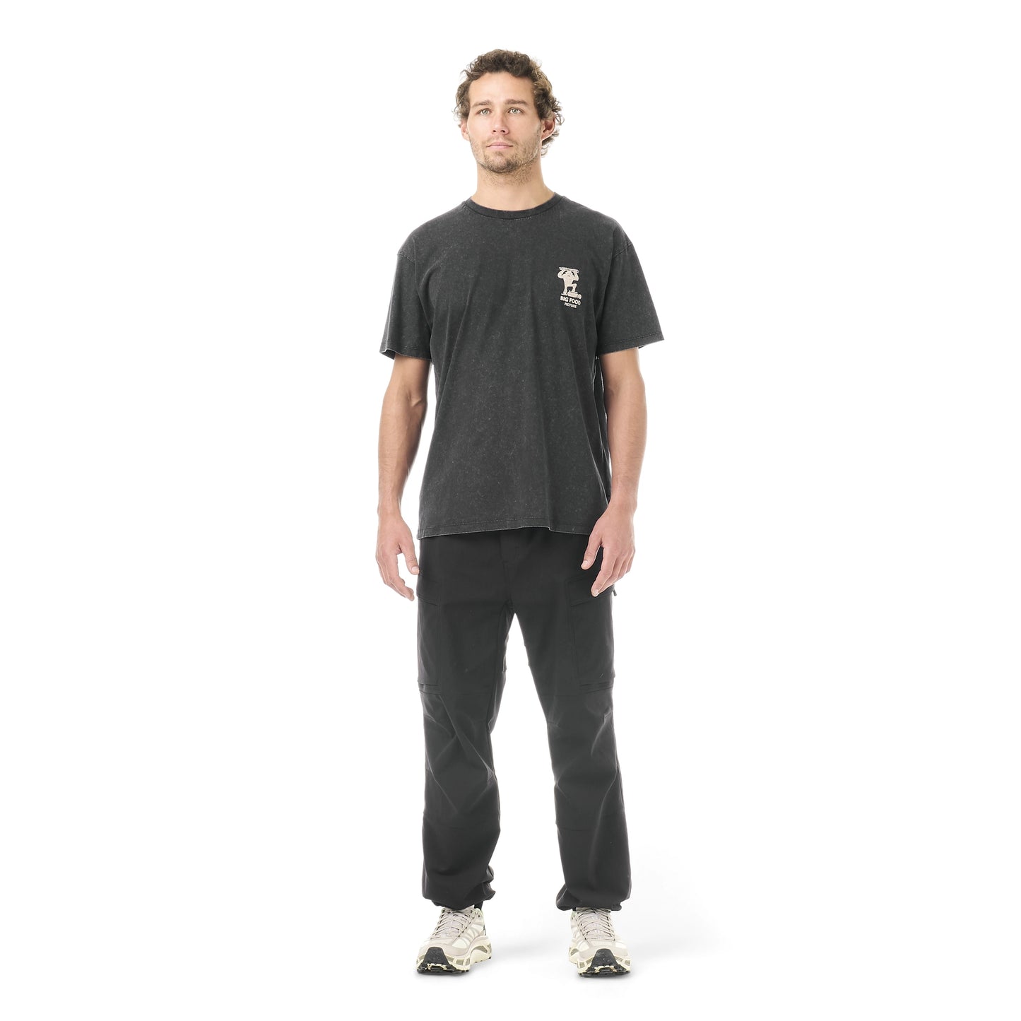 Picture Organic Big Foot Tee A man modeling a dark charcoal gray t-shirt with a small white graphic on the left chest, paired with dark gray sweatpants and white sneakers, shot against a solid white background.