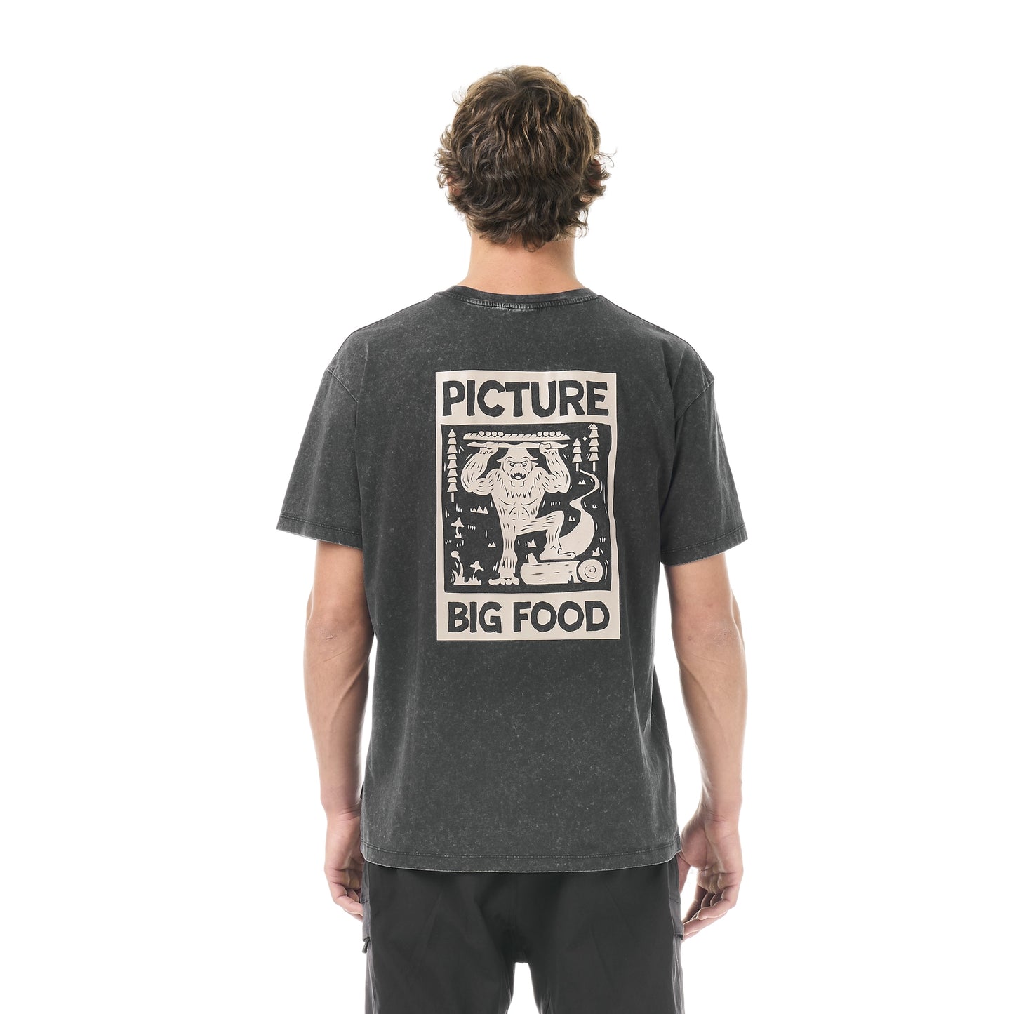 Picture Organic Big Foot Tee A man viewed from behind wearing a dark gray washed graphic t-shirt with a large cream-colored 'Picture Big Food' print on the back, paired with black pants, against a plain white background.
