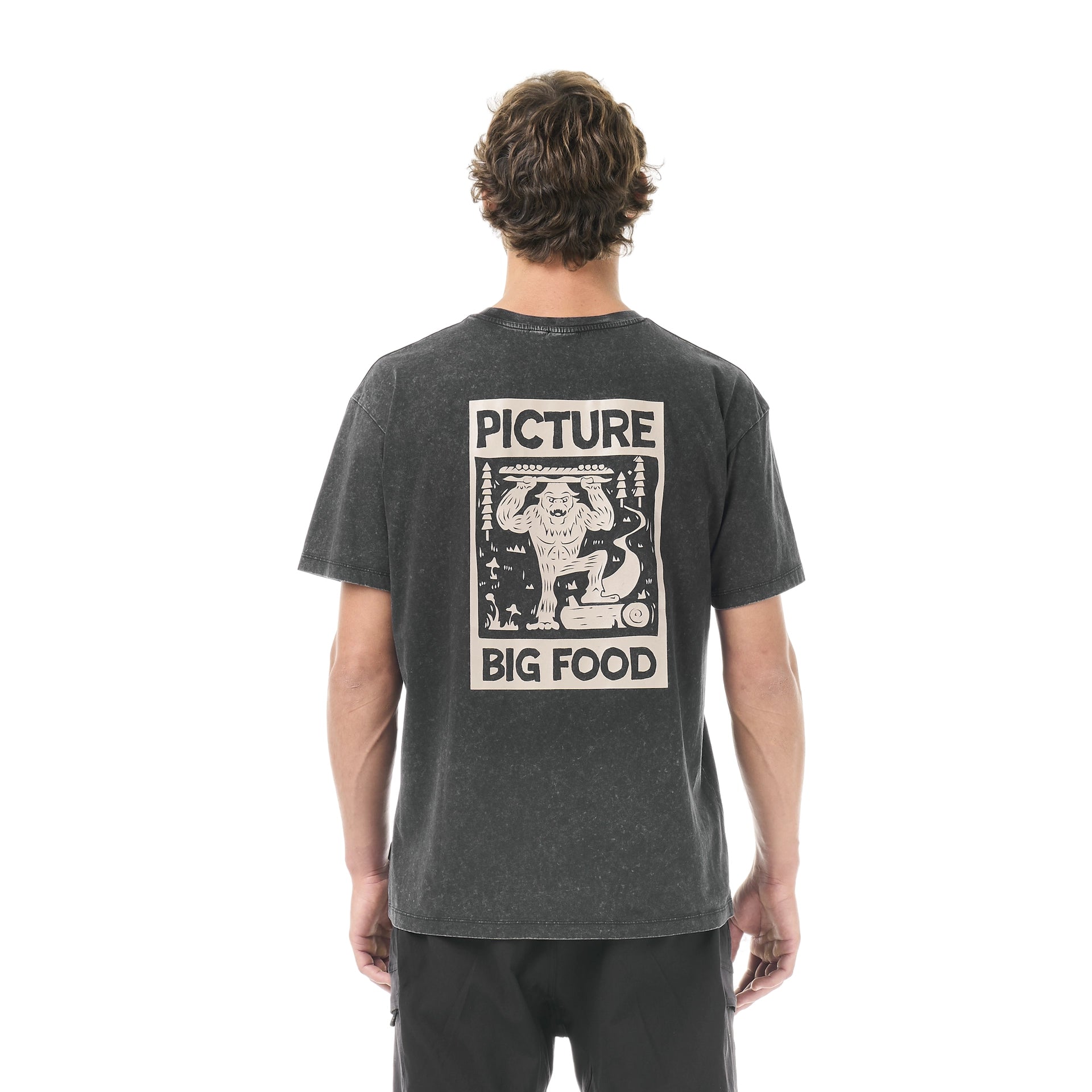 Picture Organic Big Foot Tee A man viewed from behind wearing a dark gray washed graphic t-shirt with a large cream-colored 'Picture Big Food' print on the back, paired with black pants, against a plain white background.