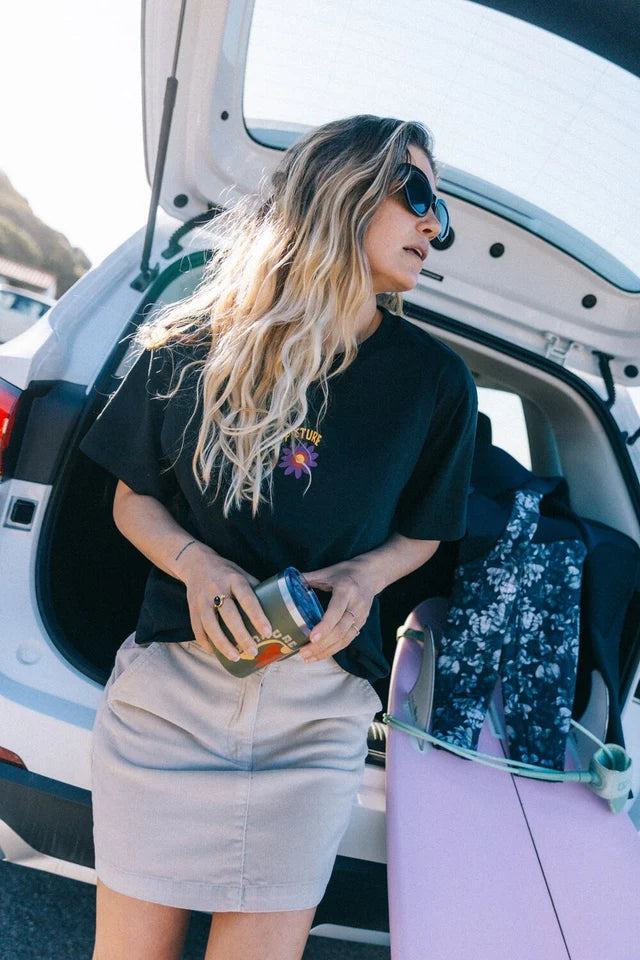 A woman stands at the back of her car with the trunk open wearing a black t shirt and cord mini skirt