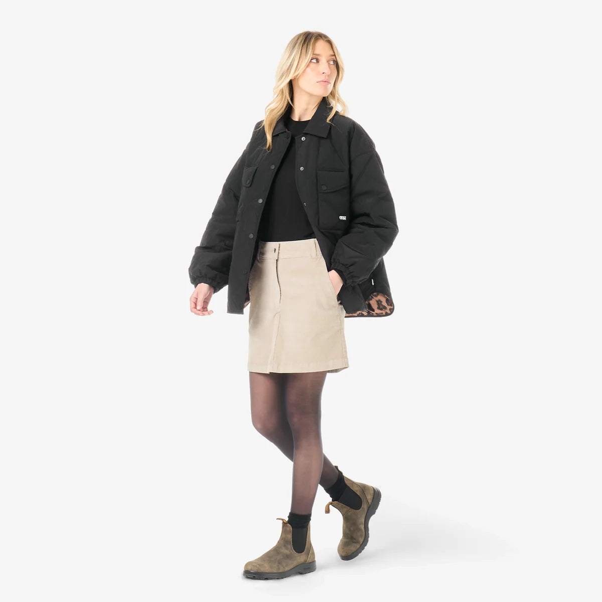 A woman stands in a white studio space wearing  black jacket, tan mini skirt and brown boots