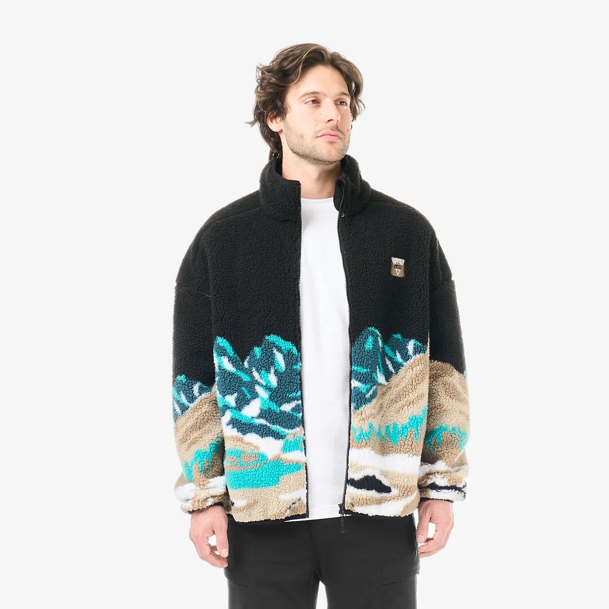 A mans stands in a white studio space wearing a black zip fleece with a mountain scape  design on the bottom half of the sweater, and black pants